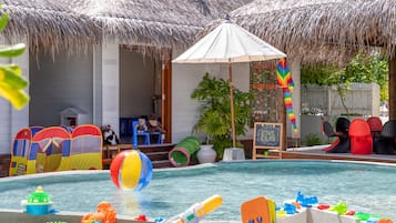 Un área de piscina con una estructura de techo de paja, juguetes inflables y un letrero del club infantil.