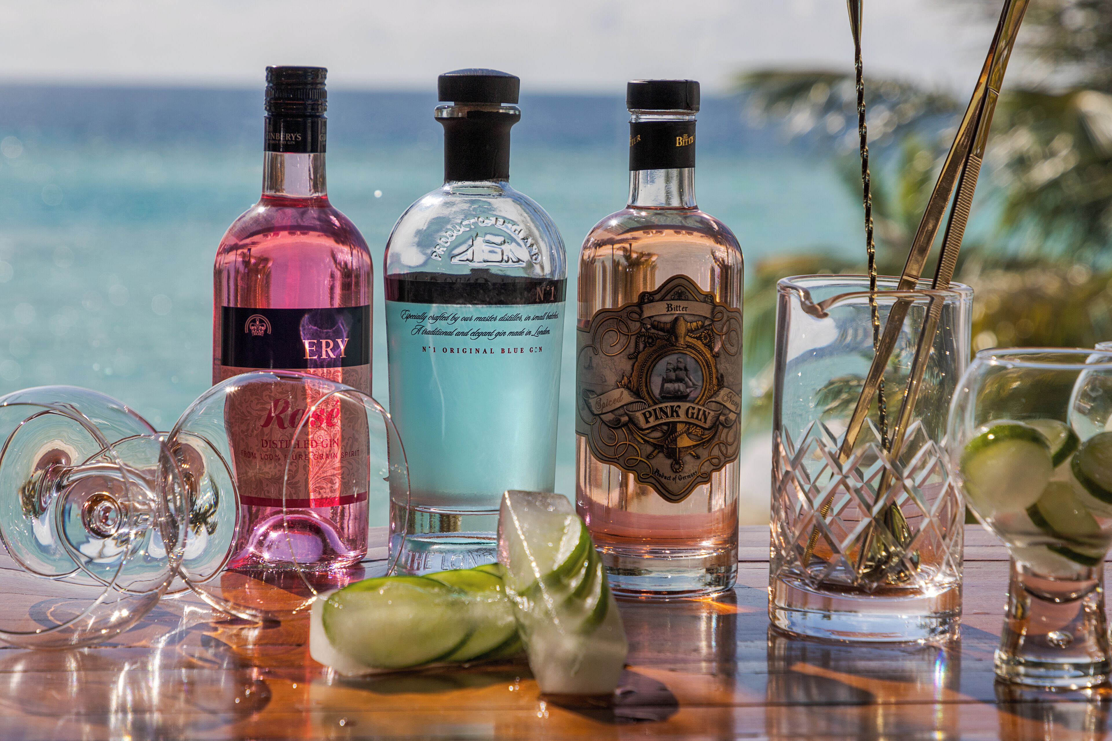 Three bottles of gin and two glasses on a table with a view of the ocean.