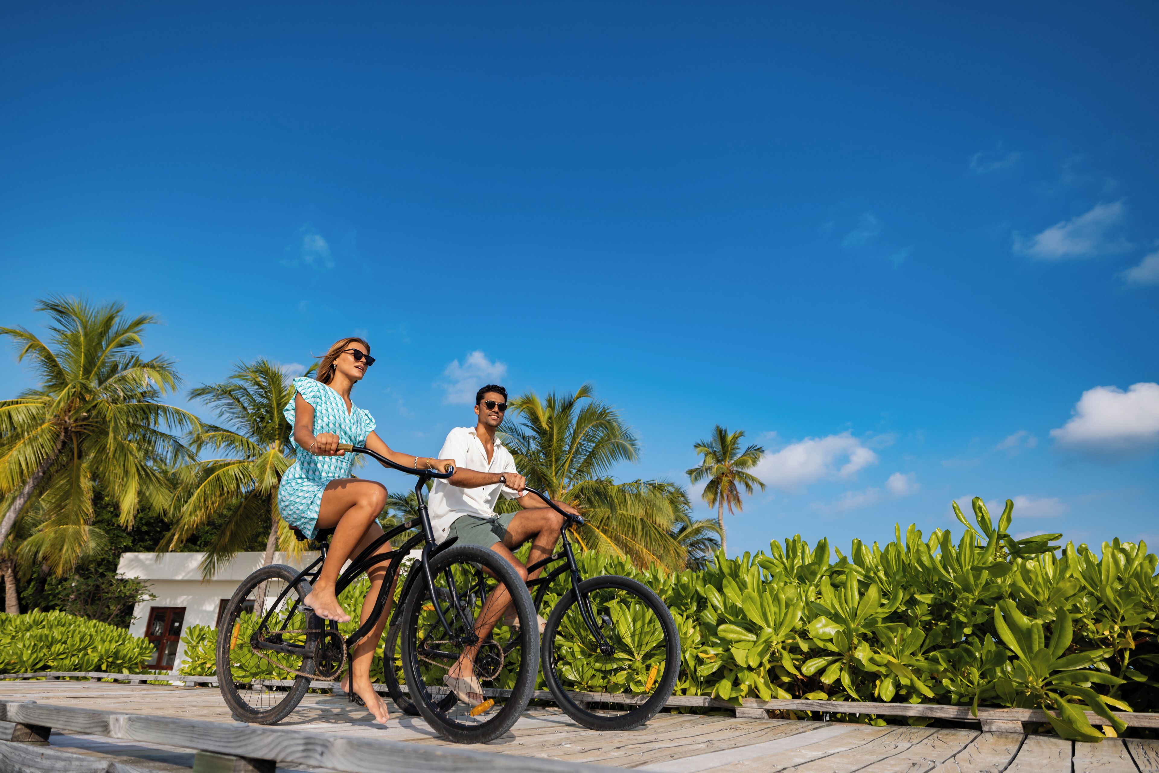 Two people riding a bicycle on a wooden deck with palm trees and greenery in the background.