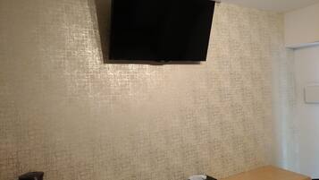 A room with a television mounted on the wall, a table with a tissue box and a remote control, and a kettle on the floor.