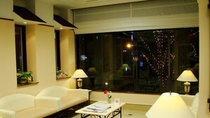 A hotel room with a sofa, a coffee table, two lamps, a window with a view of the city, and a small table with a flower vase. - West Inn Fuji-Yoshida (Fujiyoshida)