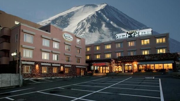 A hotel with a mountain backdrop, featuring a parking lot and multiple buildings. - West Inn Fuji-Yoshida (Fujiyoshida)