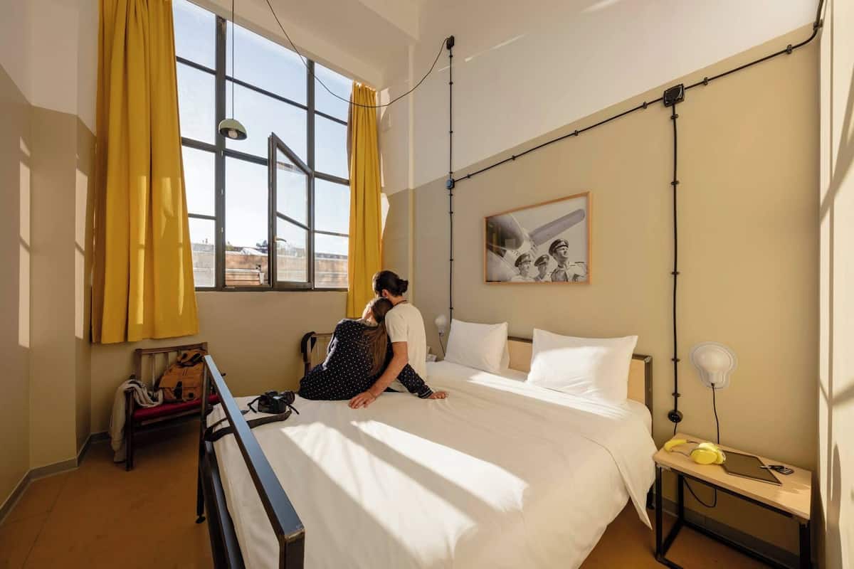 Ein Hotelzimmer mit einem Bett, einem Nachttisch mit Lampe, einem Fenster mit Vorhängen und einem an der Wand hängenden Bild.