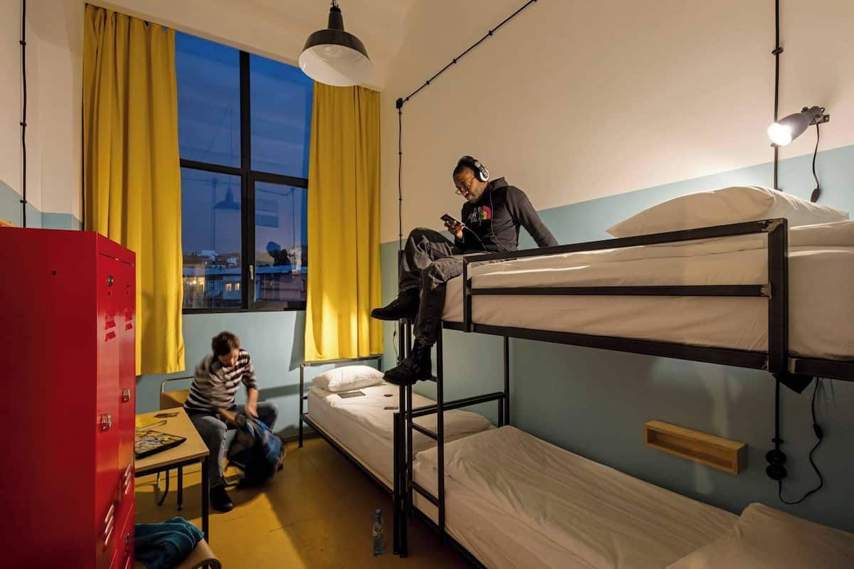 Ein Schlafsaalzimmer mit Etagenbetten, einem Schreibtisch, einem Stuhl, einem Fenster mit Vorhängen und einem roten Stauraum.