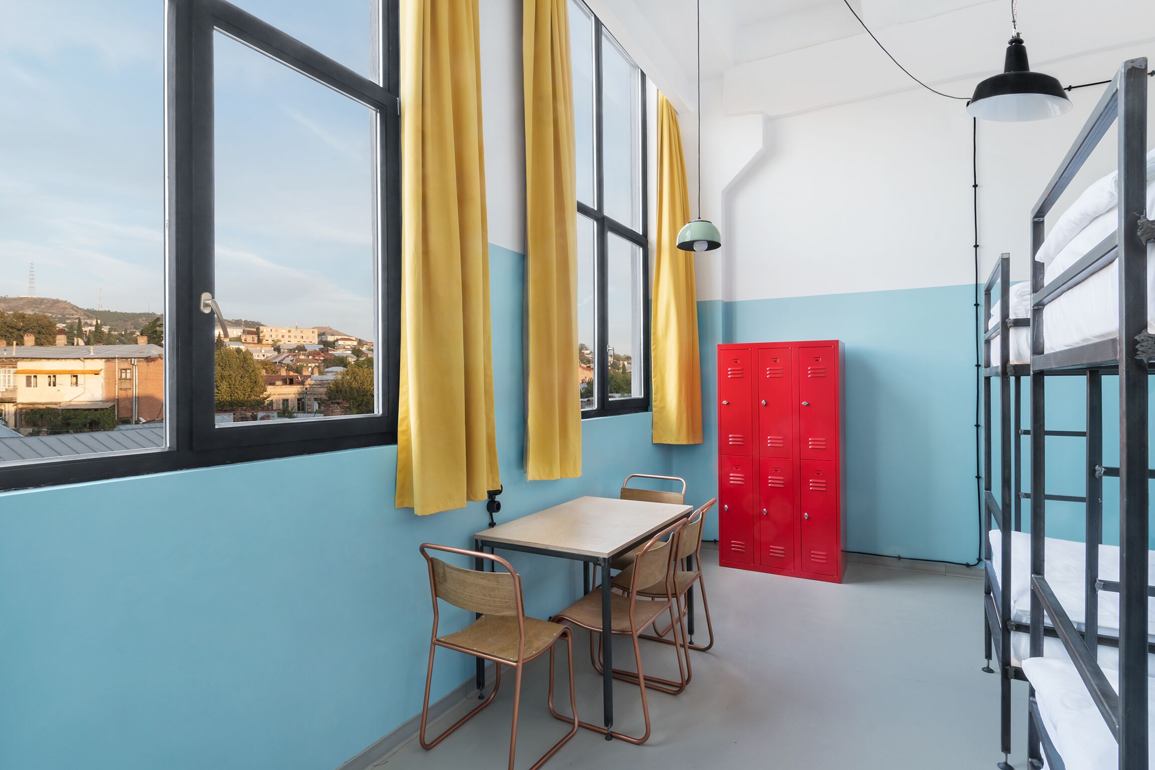 A room with a table and chairs, a red storage unit, bunk beds, and a view through a window.