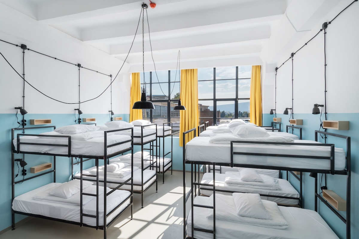 A dormitory room with bunk beds, a large window with yellow curtains, and multiple wall-mounted lamps.