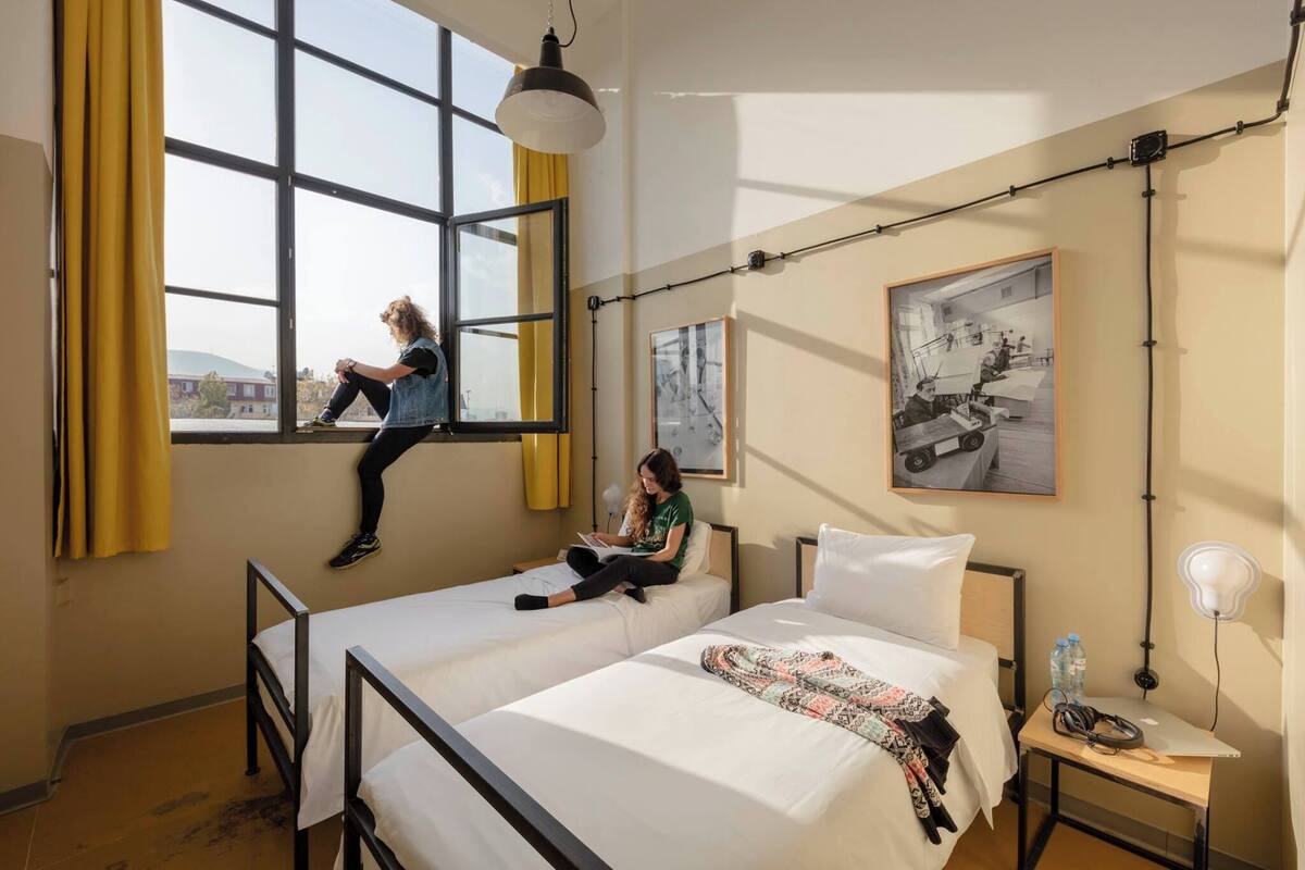 Ein Hotelzimmer mit zwei Betten, einem Fenster mit gelben Vorhängen, einem Schreibtisch mit Lampe und einem gerahmten Bild an der Wand.