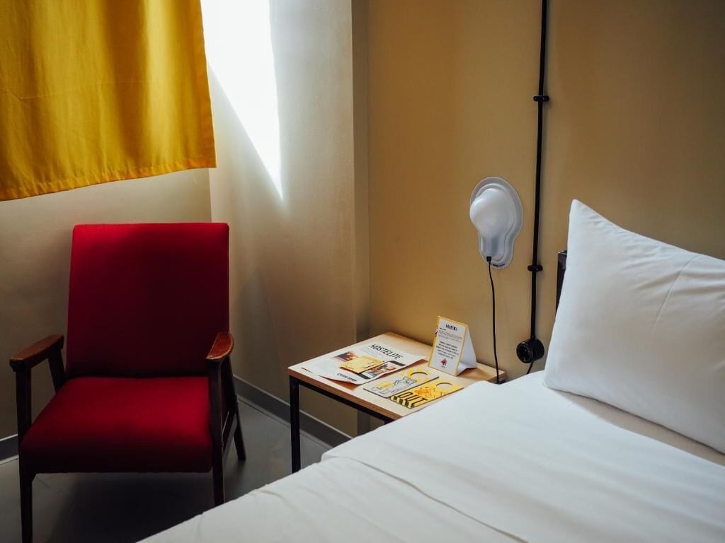 A hotel room with a bed, a red chair, a small table with a lamp, and a window with yellow curtains.
