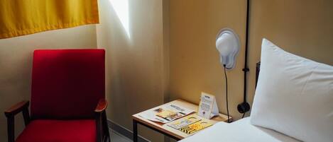 Une chambre d’hôtel avec un lit, une chaise rouge, une petite table avec une lampe et une fenêtre avec des rideaux jaunes.