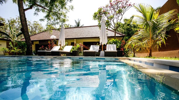 Ein Swimmingpool mit Liegestühlen und Sonnenschirmen, umgeben von tropischer Bepflanzung und einem Gebäude mit Ziegeldach.