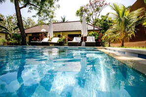 Uma piscina com espreguiçadeiras e guarda-sóis, rodeada por plantas tropicais e um edifício com telhado em azulejos.