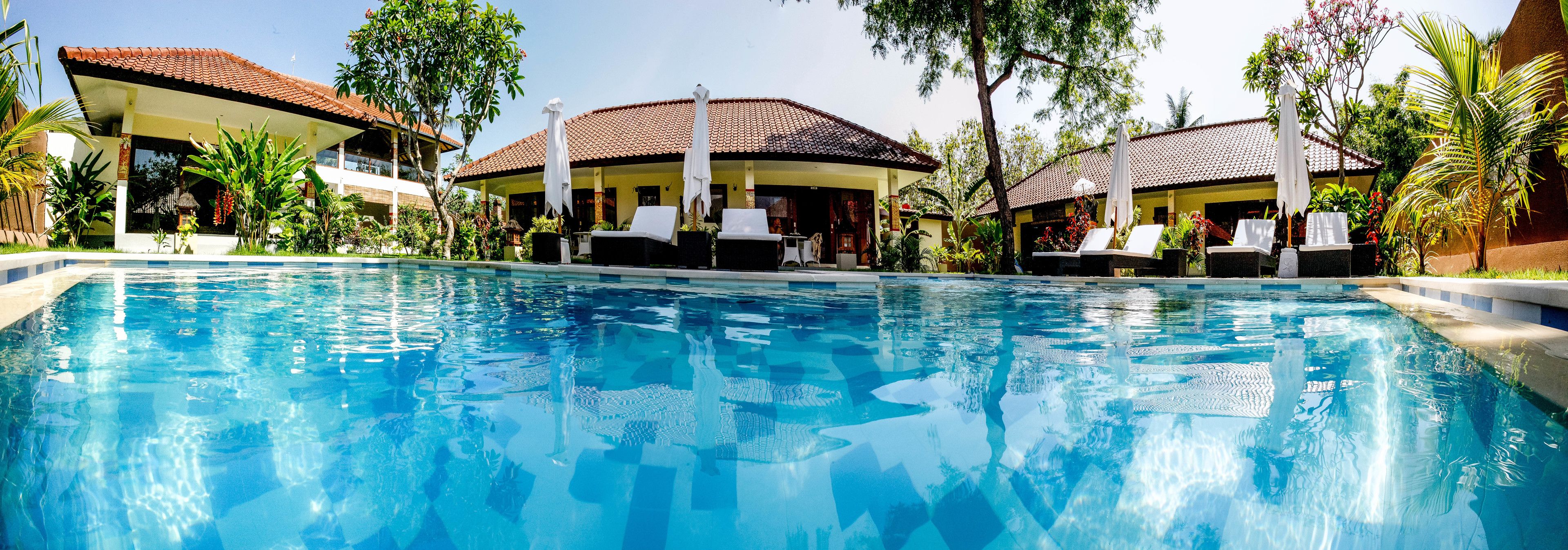 Una piscina con sillones de playa y sombrillas, rodeada de edificios con tejados de azulejos y plantas tropicales.