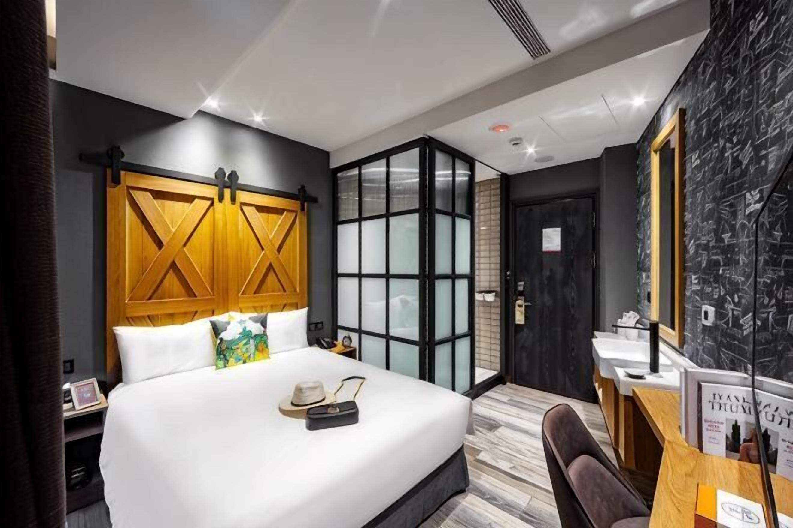 A modern hotel room with a large bed, a wooden headboard, a desk with a mirror, and a black and white patterned wall.