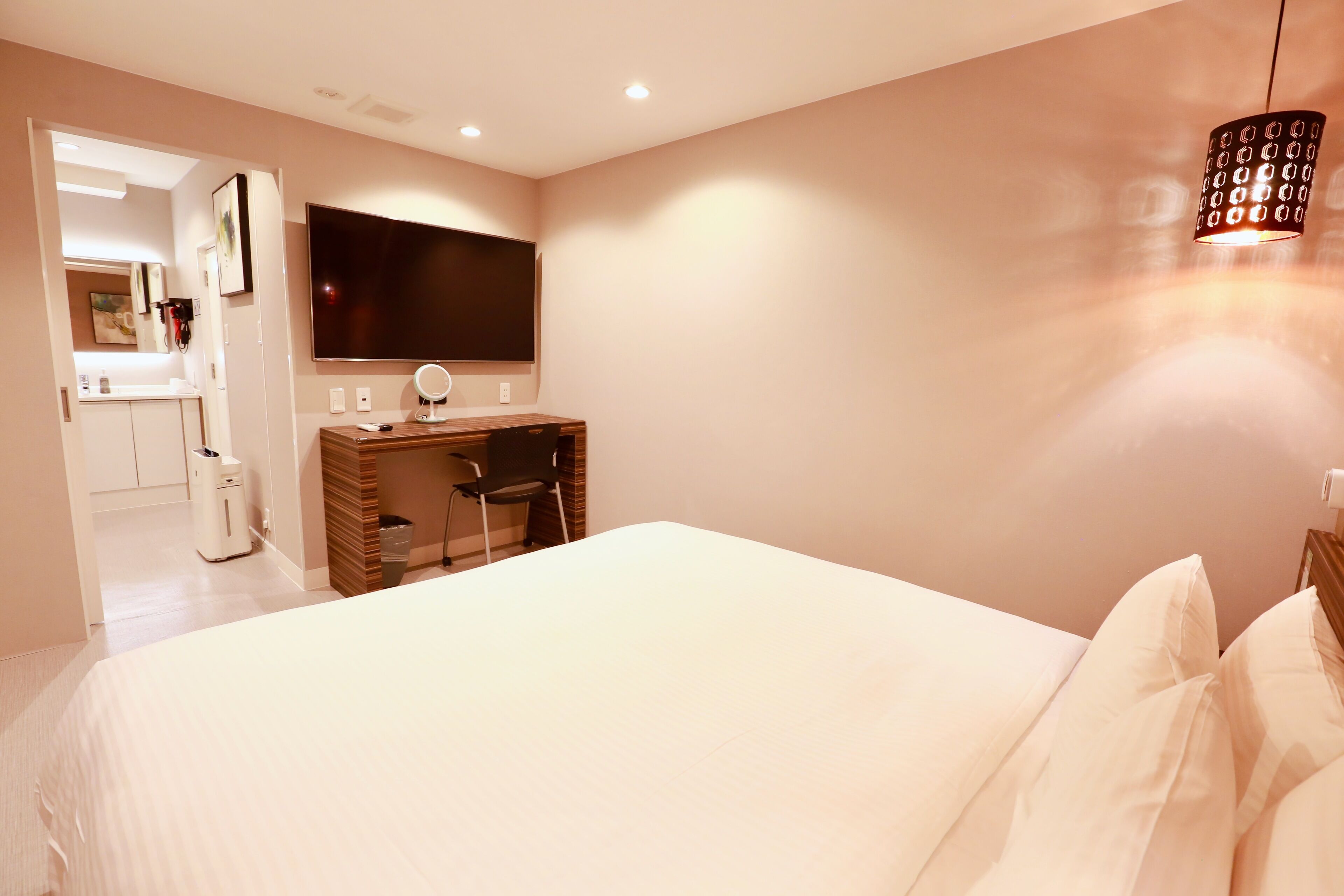 A bedroom with a large bed, a desk with a chair, a TV mounted on the wall, and a hanging light fixture.