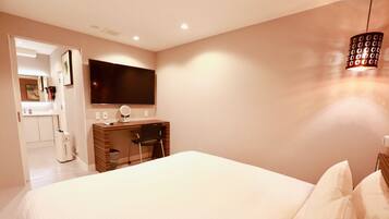 A bedroom with a large bed, a desk with a chair, a TV mounted on the wall, and a hanging light fixture.