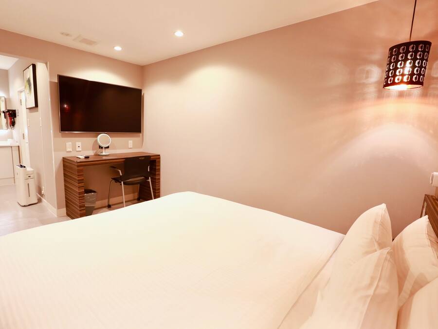 A bedroom with a large bed, a desk with a chair, a TV mounted on the wall, and a hanging light fixture.