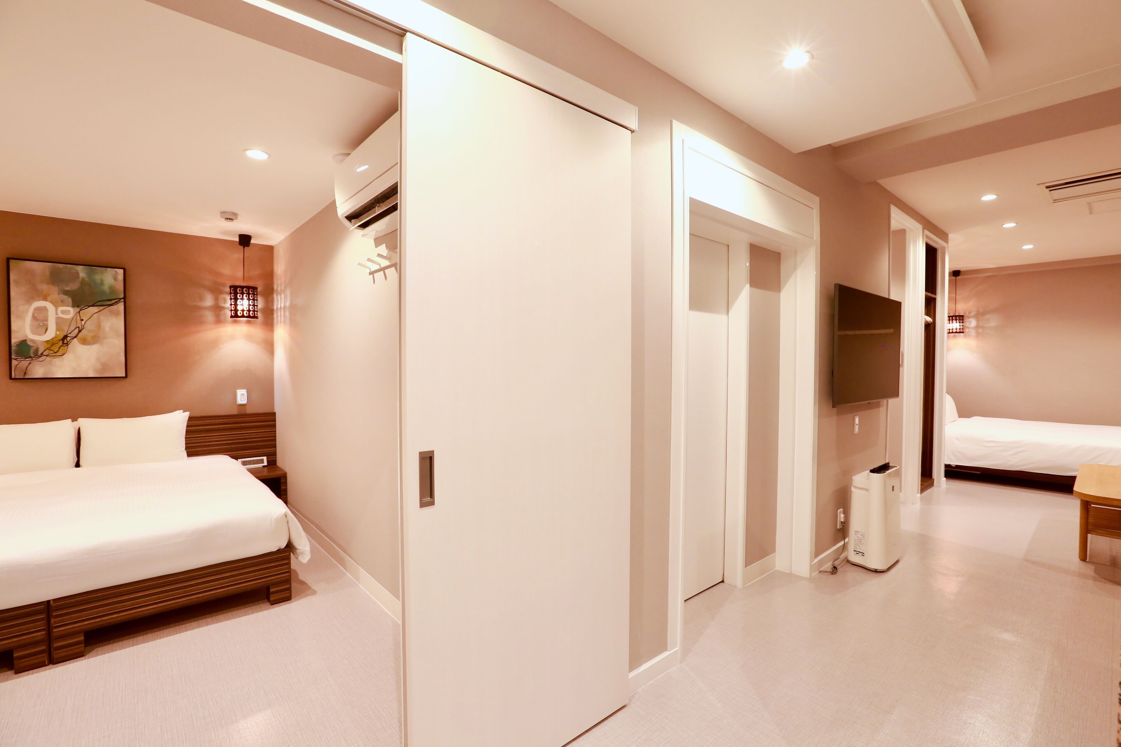 A hotel room with a bed, a bedside table, a wall-mounted TV, and a door leading to another room.