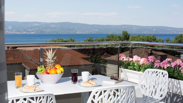Un balcón con una mesa lista para el desayuno, que incluye frutas, bollería y bebidas, con vistas a un paisaje marino y colinas al lejos.