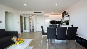 Una sala de estar moderna con un sofá negro, un comedor con cuatro sillas, una cocina con armarios blancos y un televisor grande en la pared.