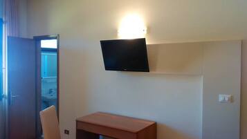 A room with a flat-screen TV mounted on the wall, a wooden desk, a chair, and a door leading to a bathroom.