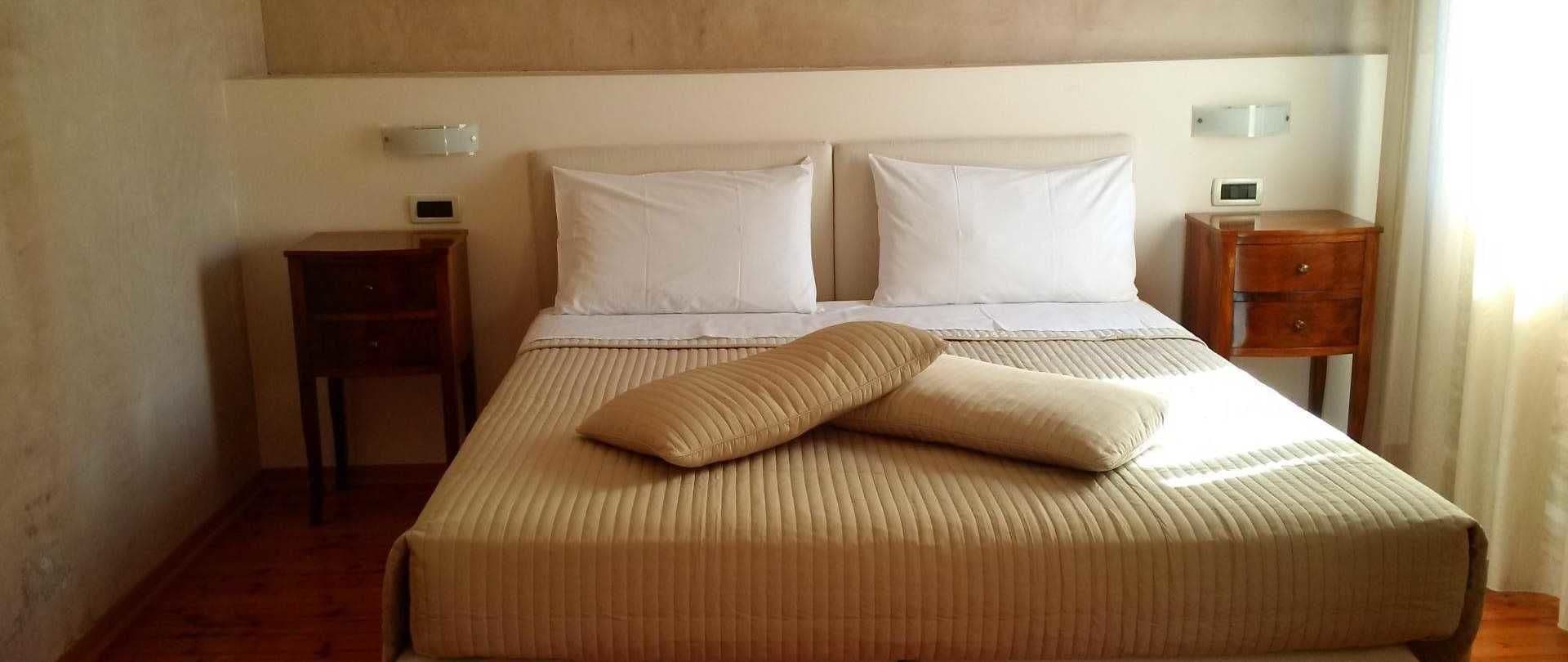 A neatly made bed with a beige cover, two white pillows, and a single beige pillow on top. There are two bedside tables, each with a small lamp and a digital clock. The room has a wooden floor and a plain wall.