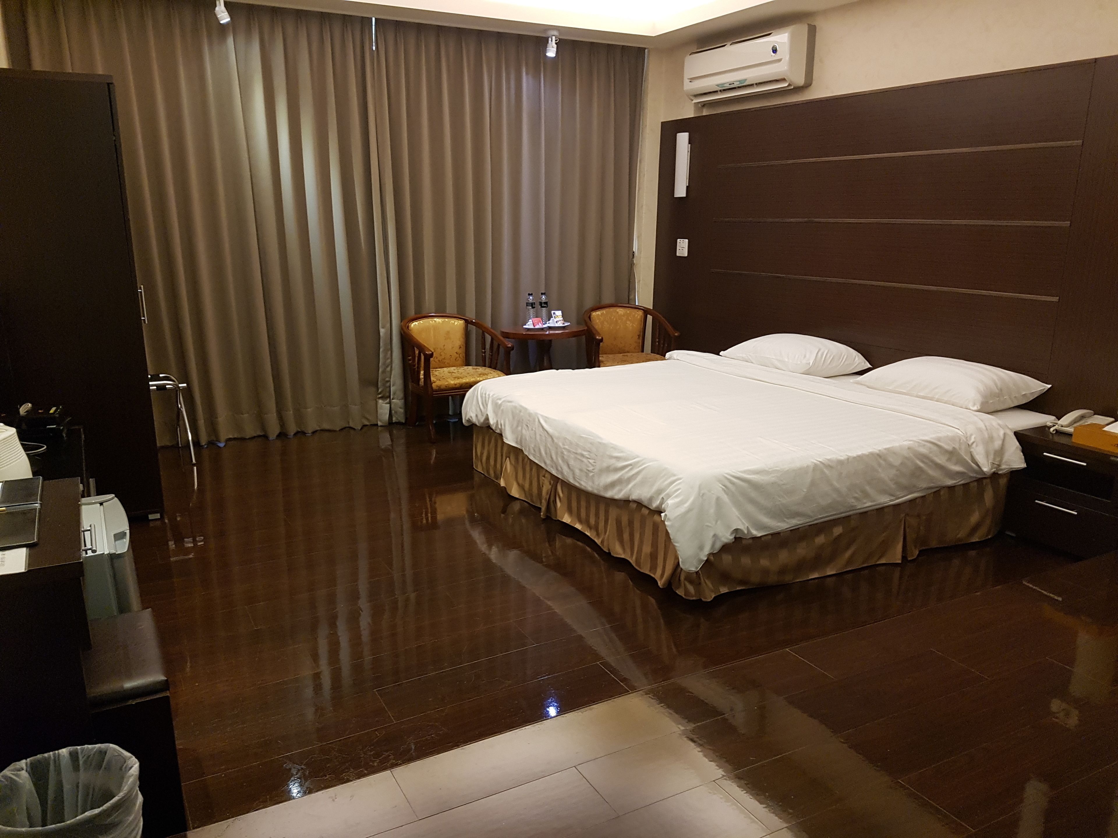 A hotel room with a large bed, two chairs, a small table, and an air conditioning unit.