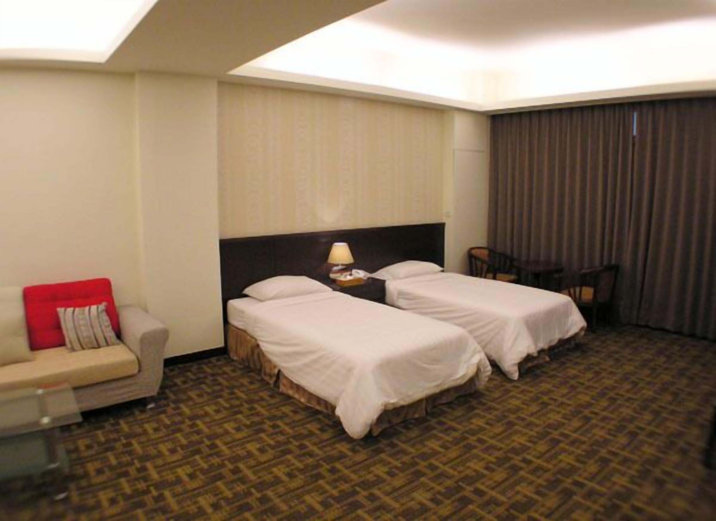 A hotel room with two single beds, a small sofa, a desk with a lamp, and a chair.