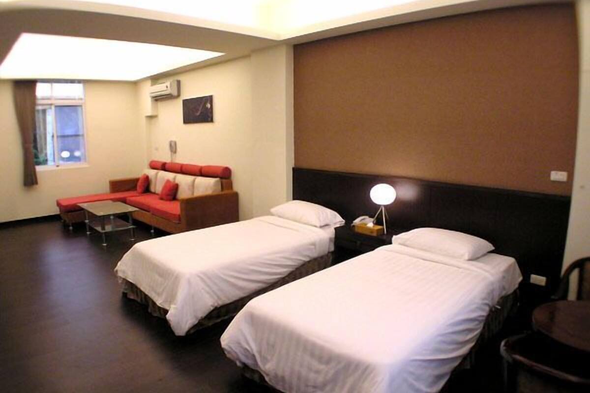 Habitación de hotel con dos camas individuales, un sofá rojo, una mesita y una lámpara.