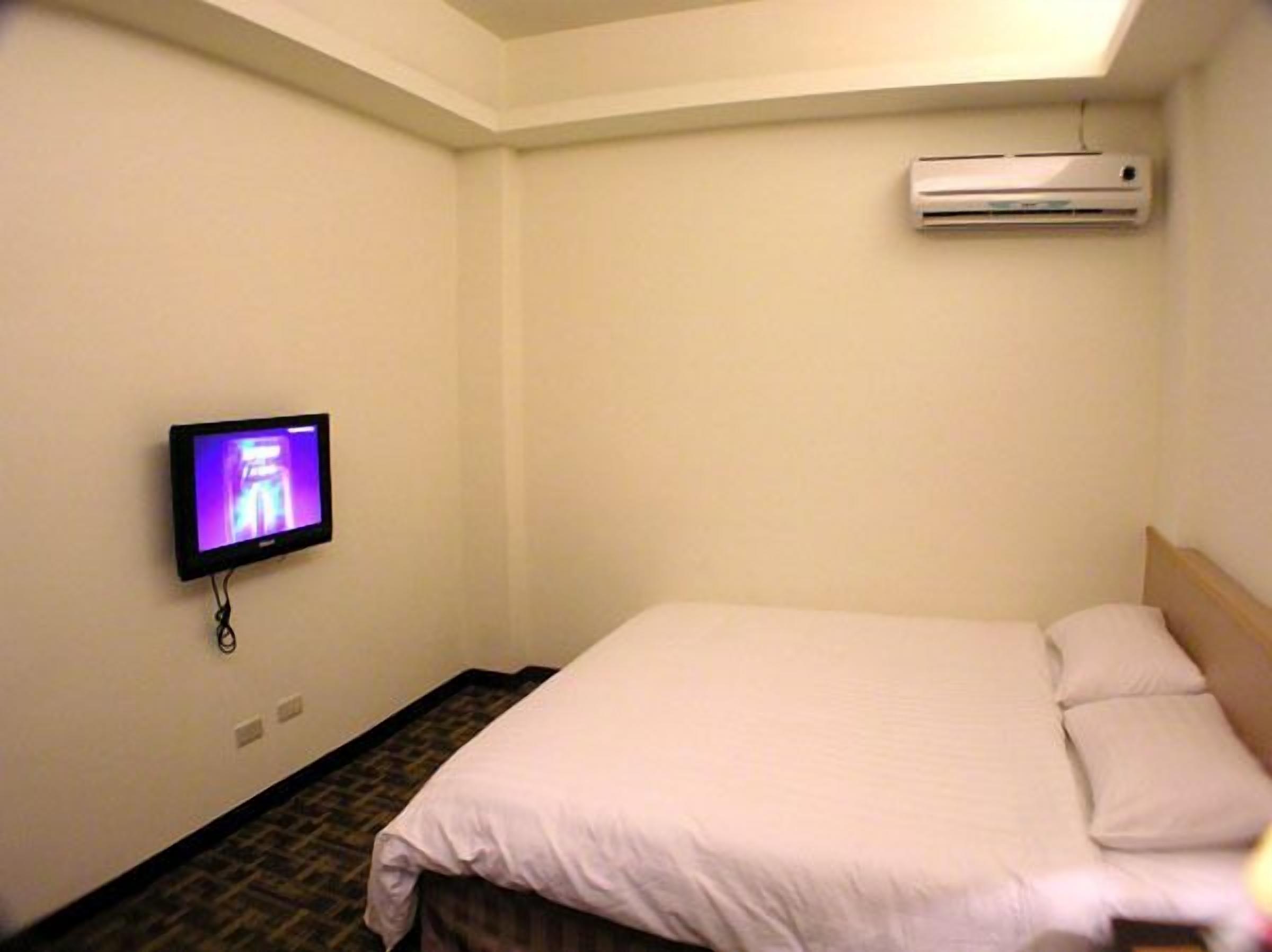 A small, simple hotel room with a bed, a television mounted on the wall, and an air conditioning unit above the bed.