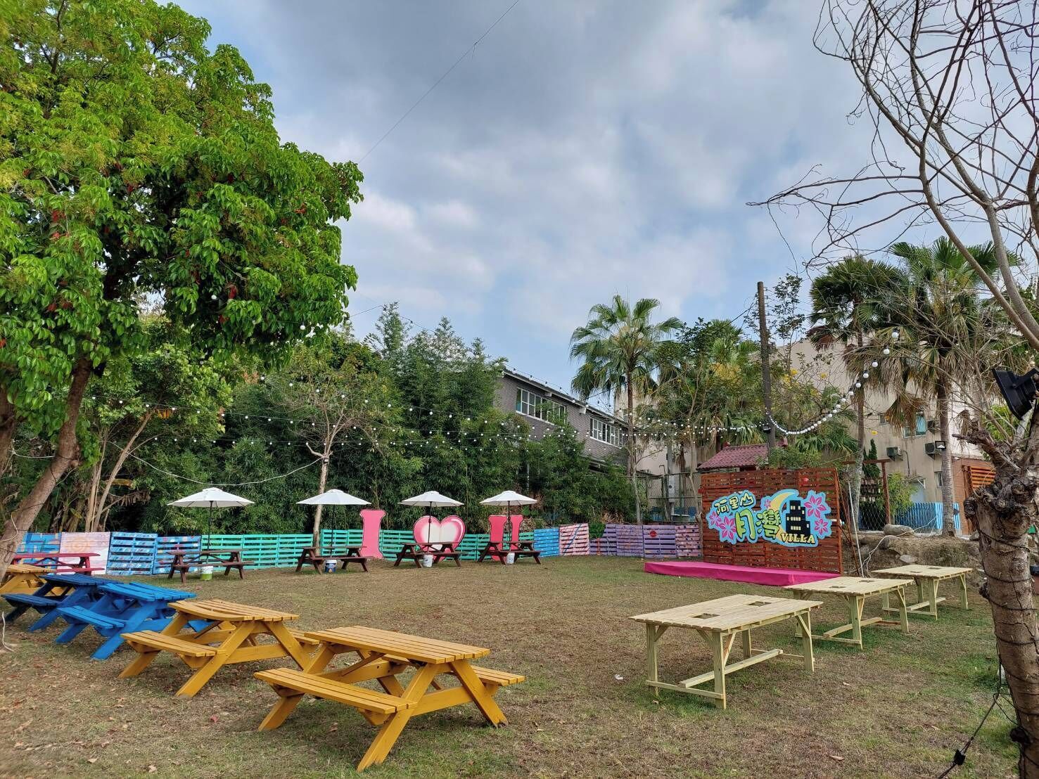 An outdoor seating area with picnic tables and umbrellas, surrounded by trees and a building in the background.