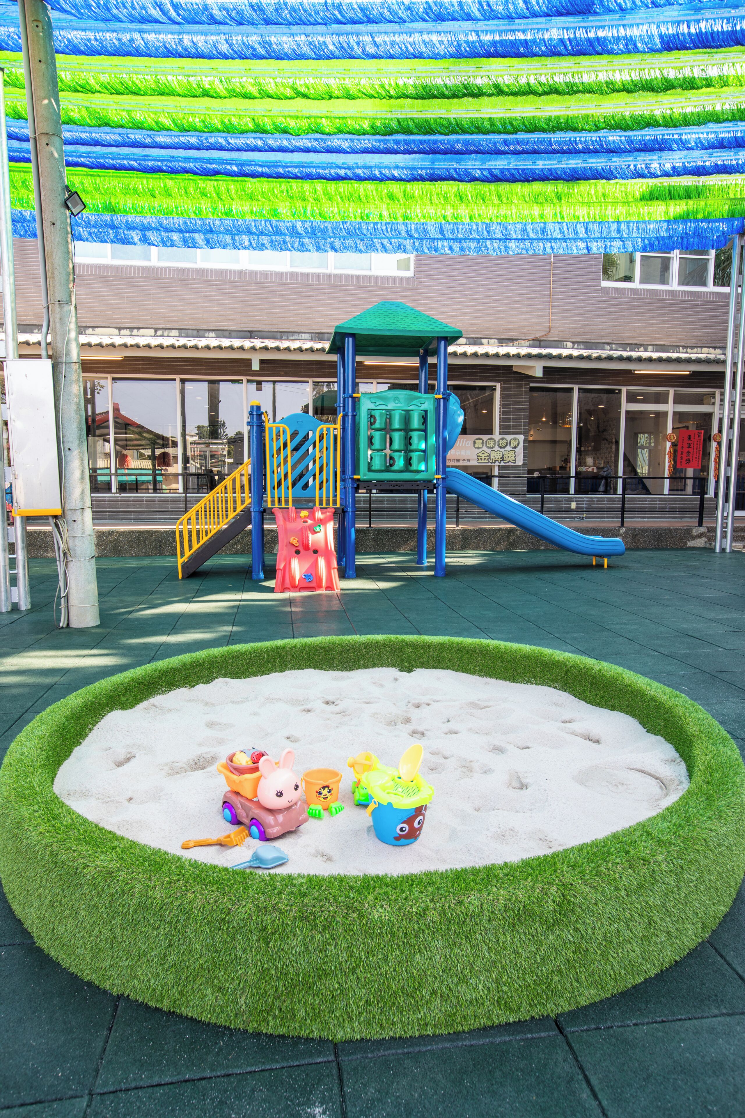 A playground with a slide and climbing structures, surrounded by a sandy play area and artificial grass.