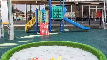 A playground with a slide and climbing structures, surrounded by a sandy play area and artificial grass.