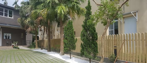 A paved courtyard with a wooden fence, palm trees, and potted plants.