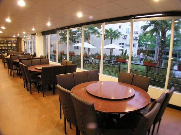 Restaurante com mesas e cadeiras redondas, grandes janelas com vista para palmeiras e edifícios.