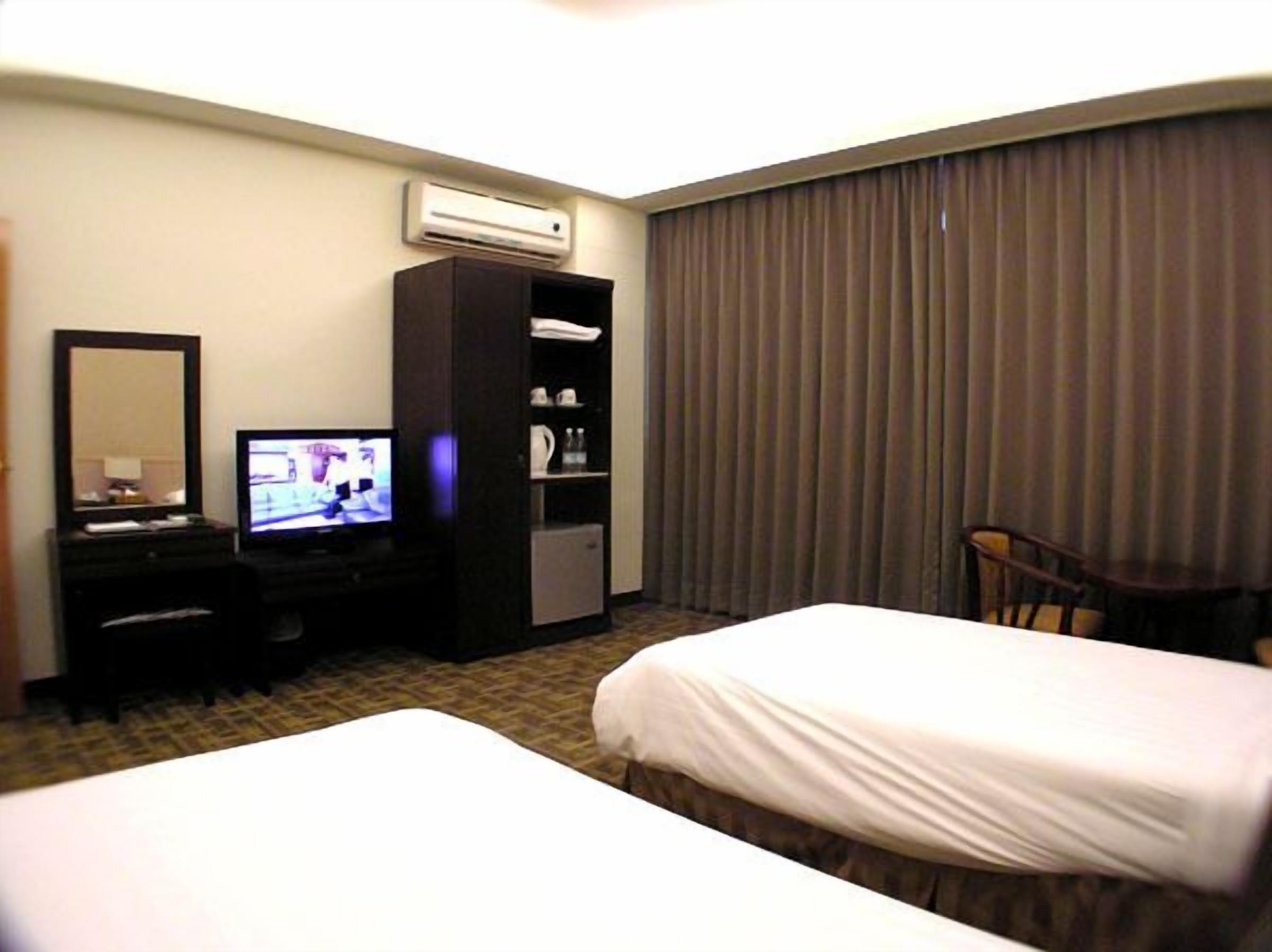 A hotel room with two beds, a TV, a minibar, and a large window with curtains.