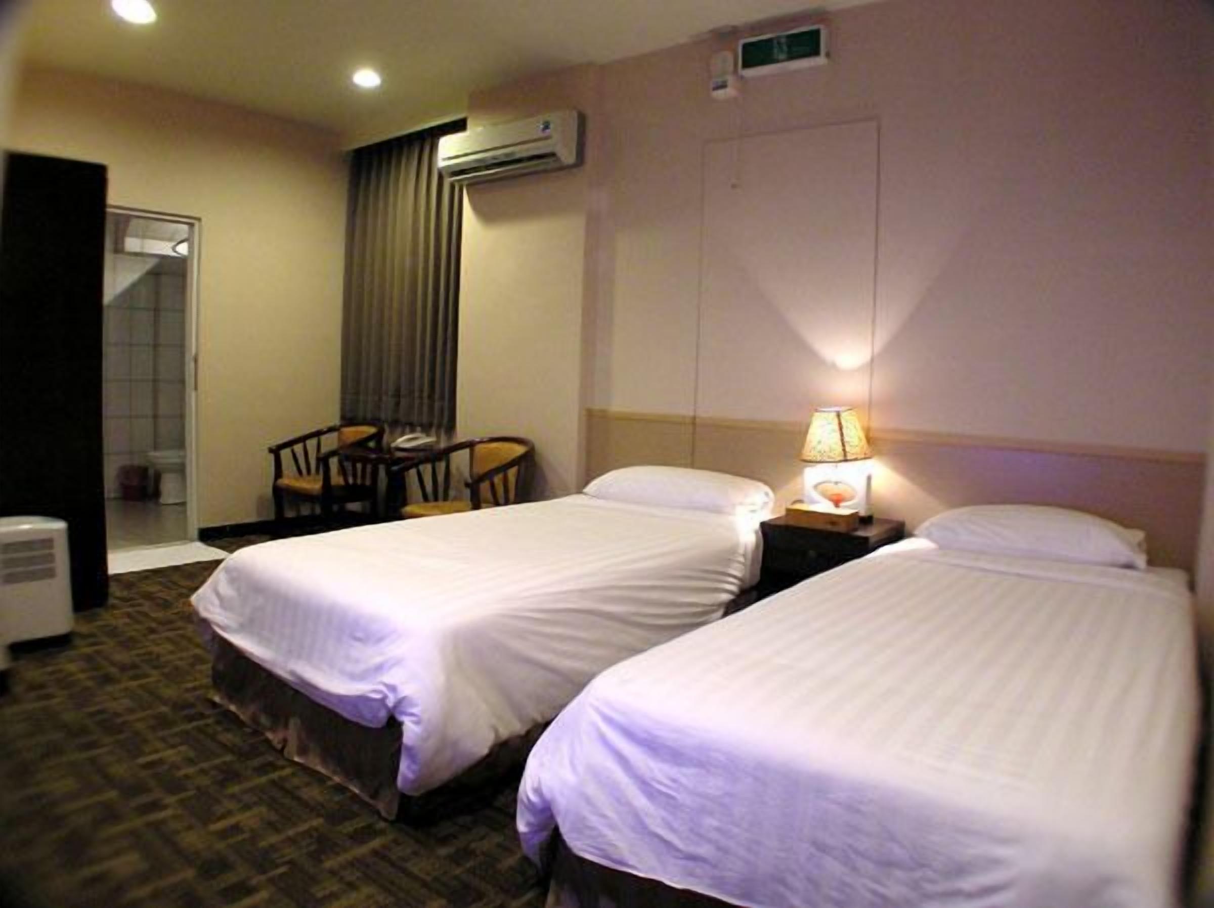 A hotel room with two single beds, a chair, a table, an air conditioning unit, and a lamp.