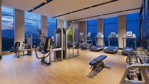 A modern gym with various exercise equipment, large windows, and a view of the cityscape.