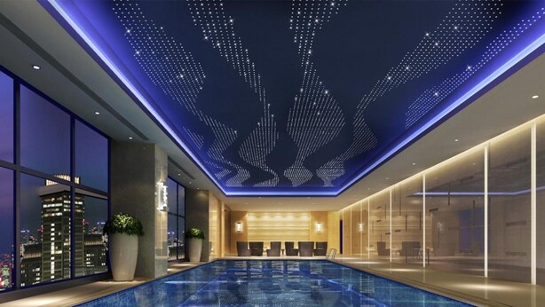 A modern indoor swimming pool with a digital ceiling display, large windows, and a city view.