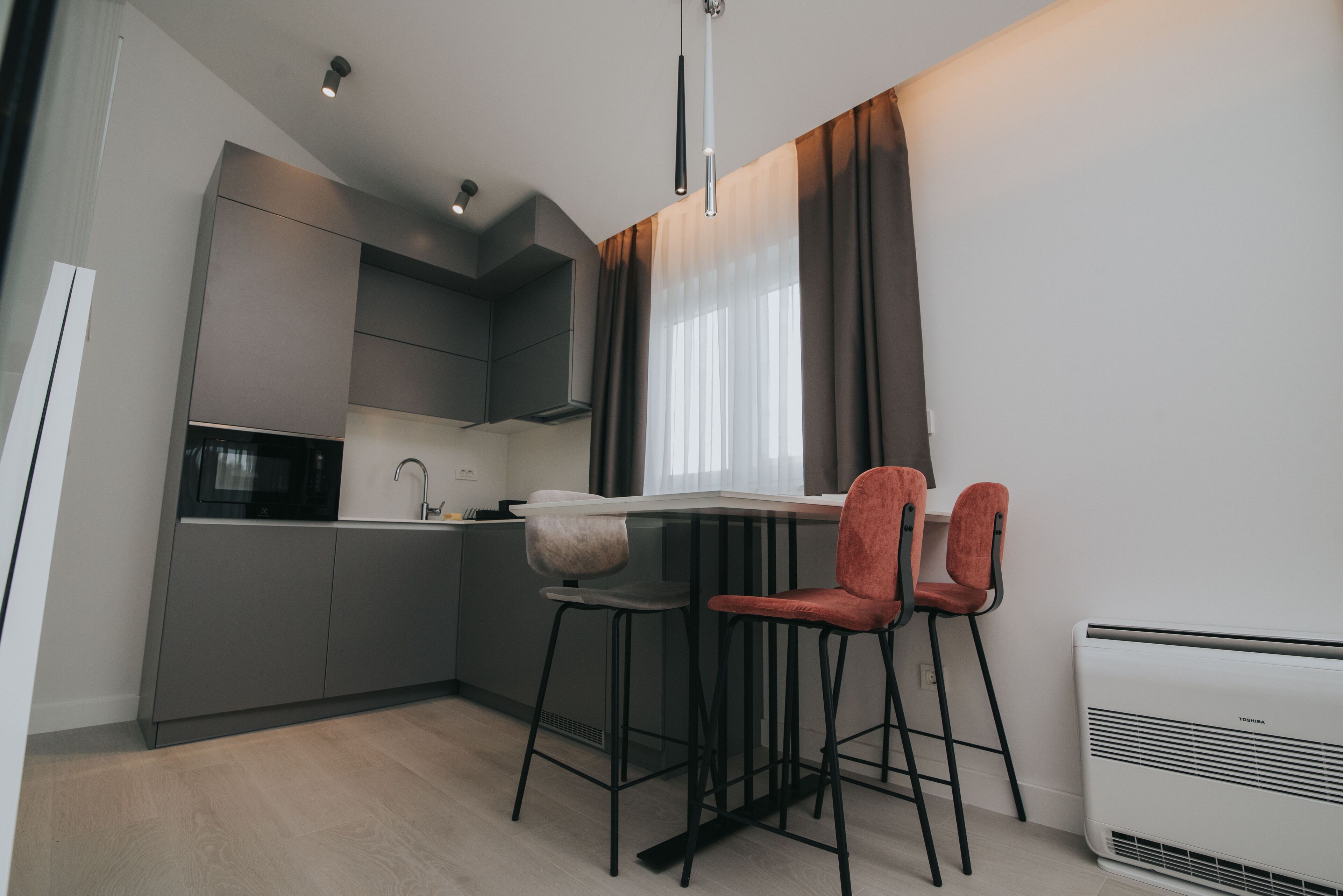 Une cuisine moderne avec des meubles gris, un micro-ondes noir et un plan de travail blanc. On y trouve deux chaises rouges et une table blanche.