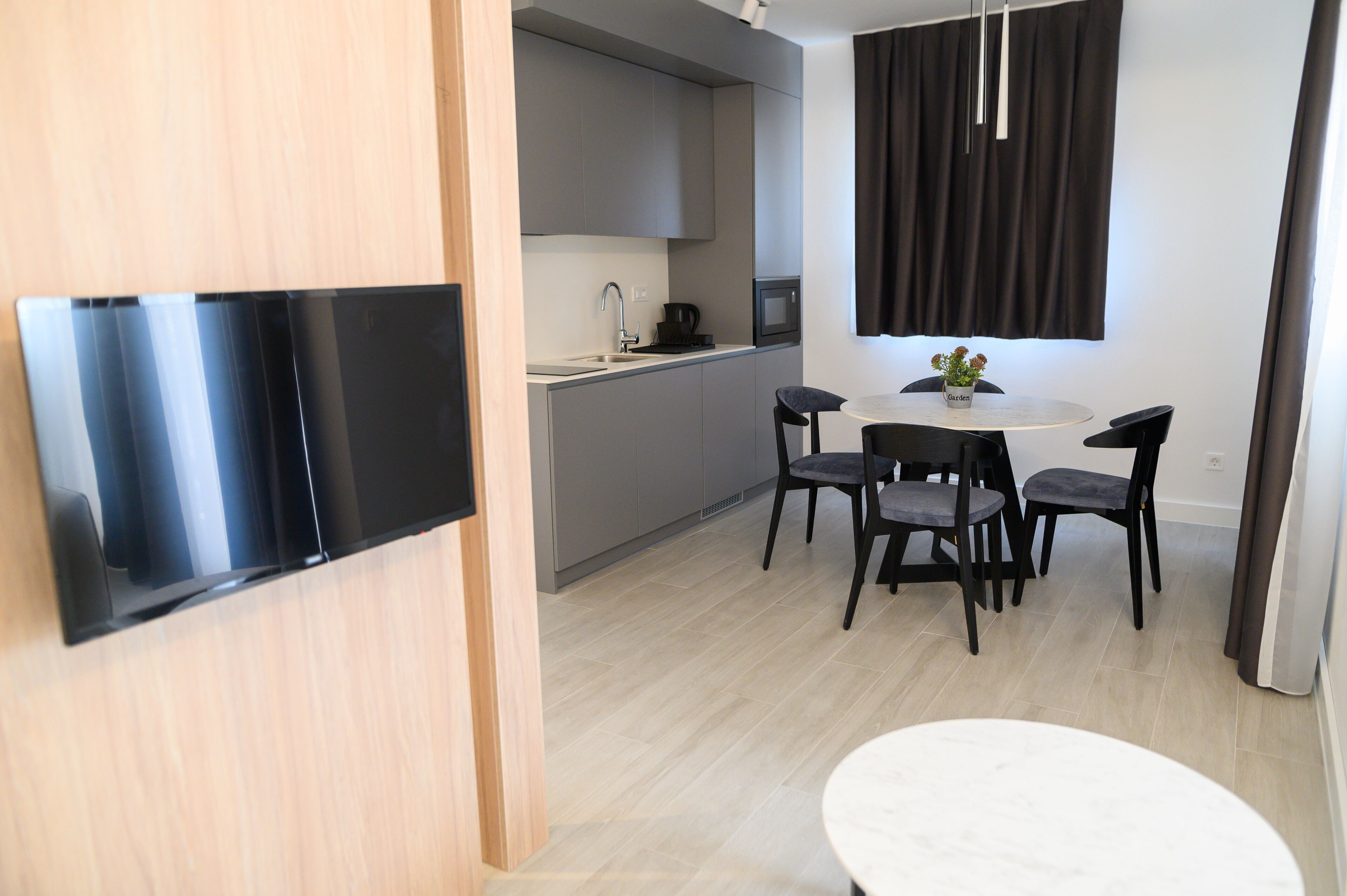 A modern kitchen with grey cabinets, a black countertop, and a microwave. There is a dining area with a round table and four chairs. A flat-screen TV is mounted on the wall.