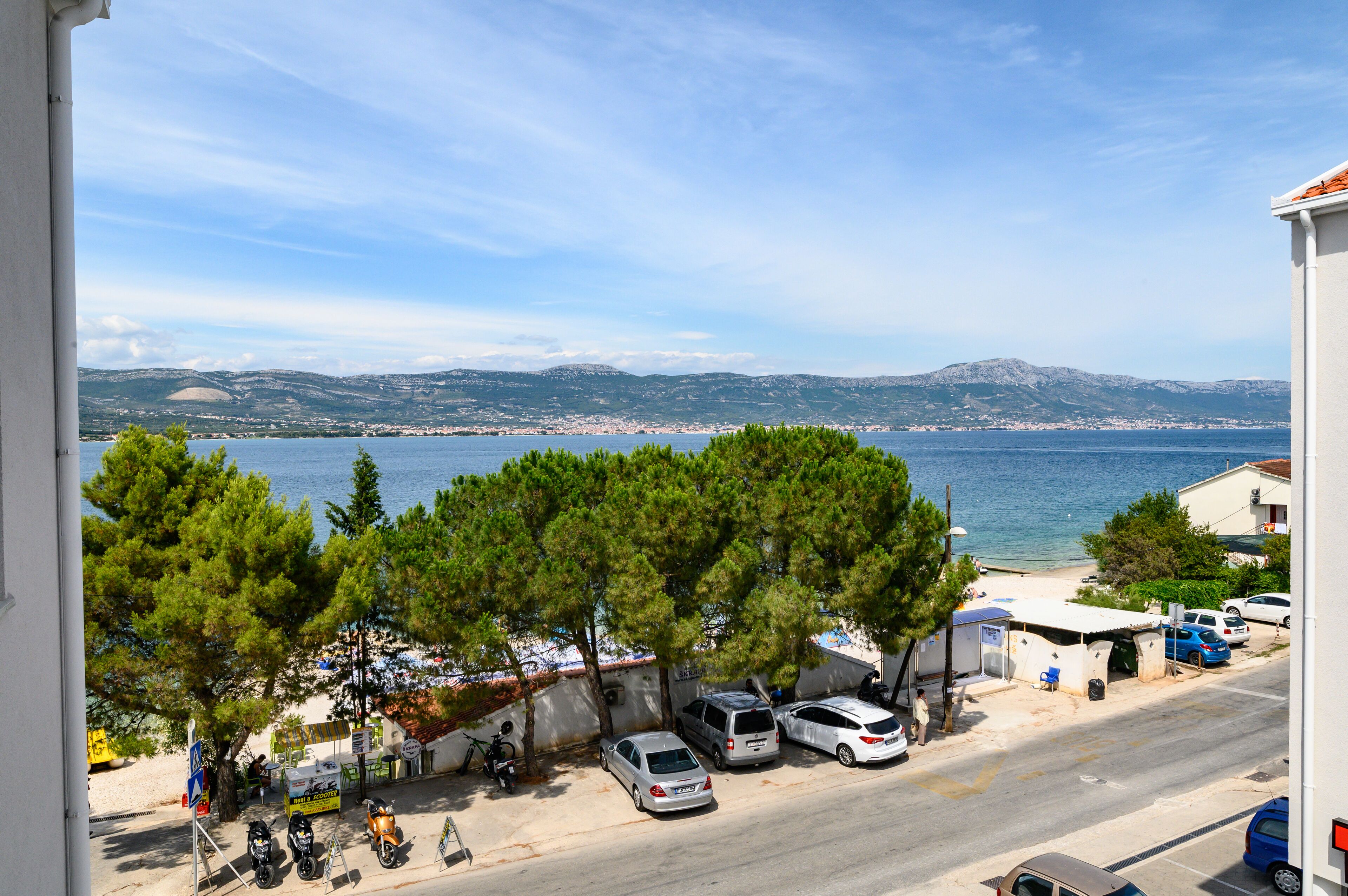 A coastal street with parked cars, a small kiosk, and a view of the sea and mountains.