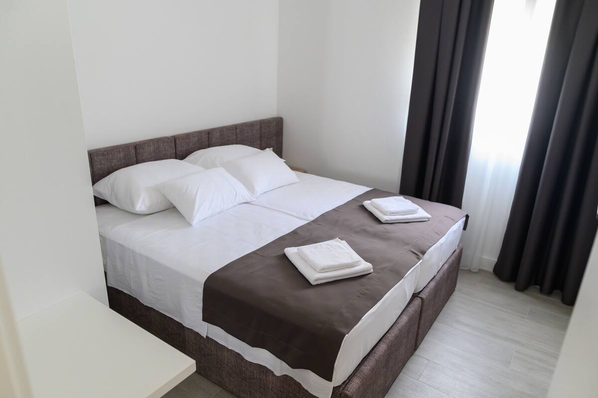 A neatly made bed with white and brown linens, a brown headboard, and a window with dark curtains.