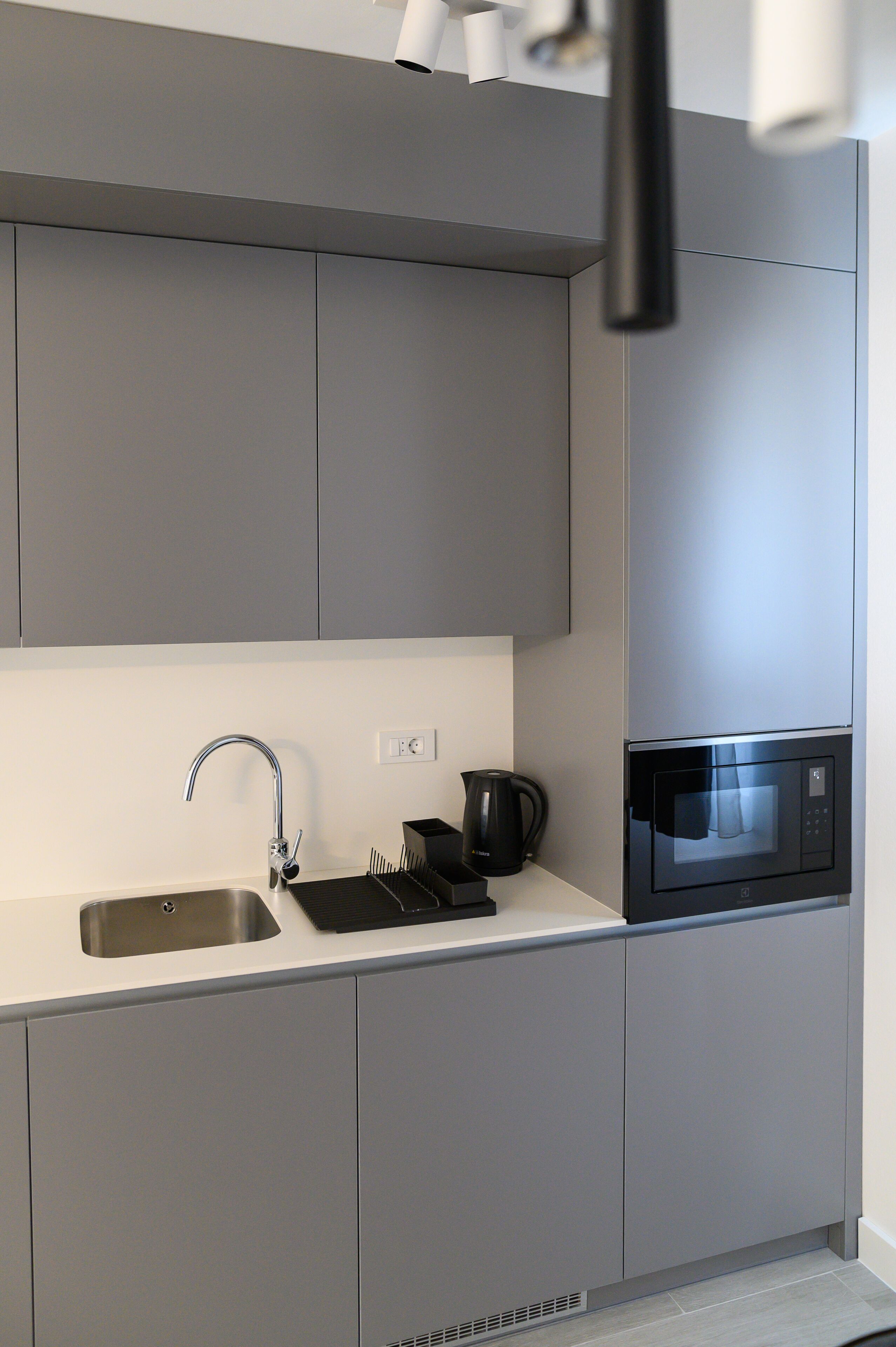 Une cuisine moderne avec des meubles gris, un évier en acier inoxydable, une bouilloire et un four à micro-ondes encastré.