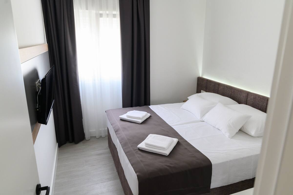 A hotel room with a bed, a flat-screen TV mounted on the wall, and a window with curtains.