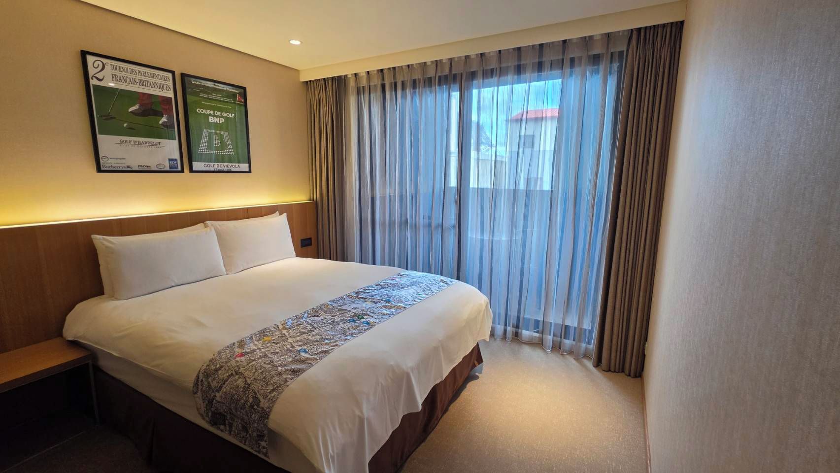 Habitación de hotel con una cama grande, una mesita de noche, un ventanal con cortinas translúcidas y dos carteles enmarcados en la pared.