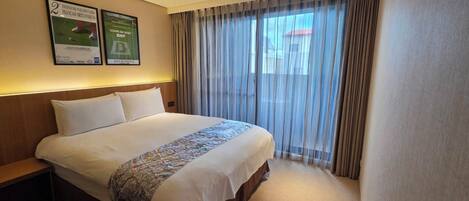 Habitación de hotel con una cama grande, una mesita de noche, un ventanal con cortinas translúcidas y dos carteles enmarcados en la pared.
