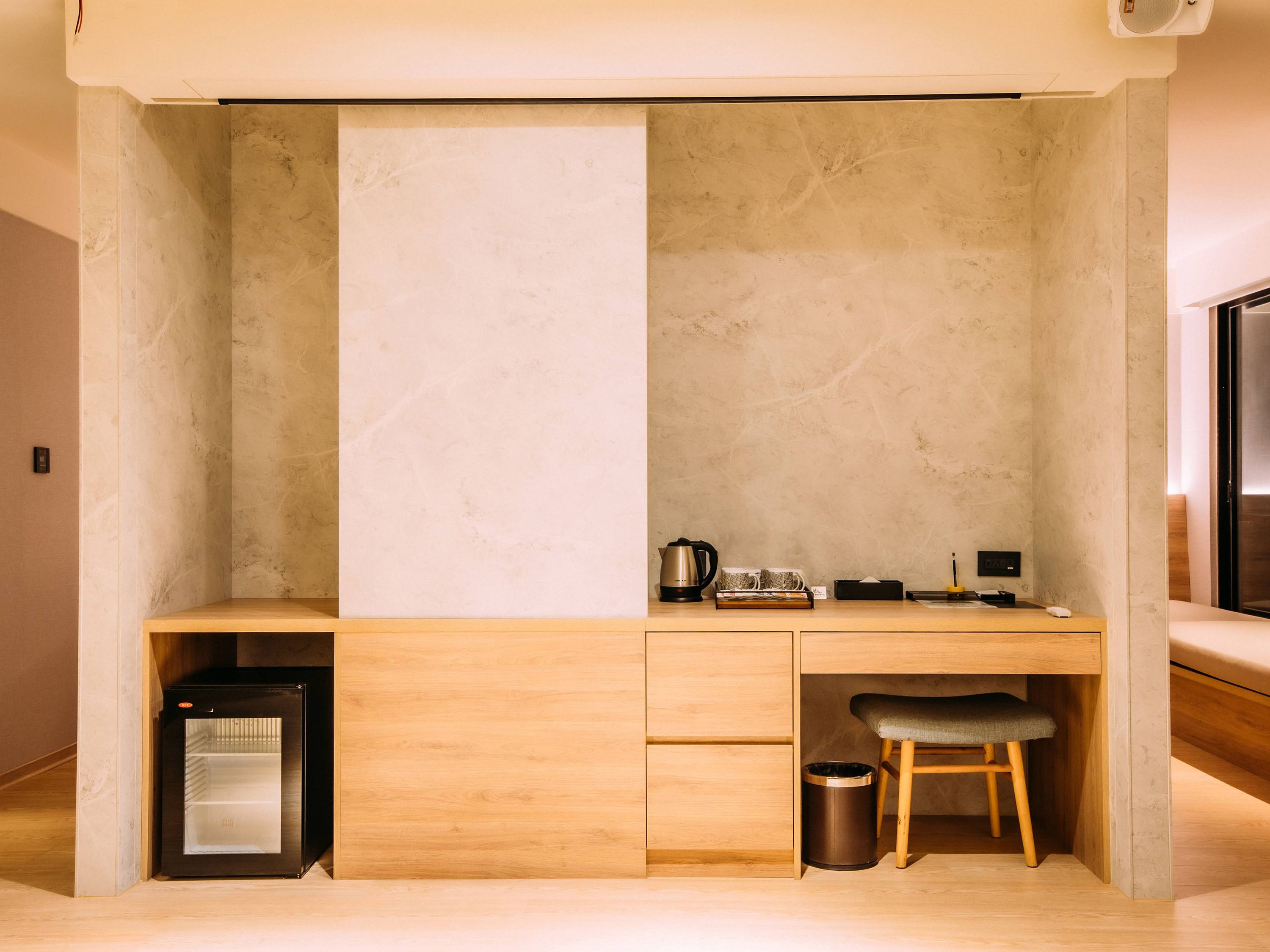 A compact room with a built-in desk, a small refrigerator, a trash bin, and a bench.