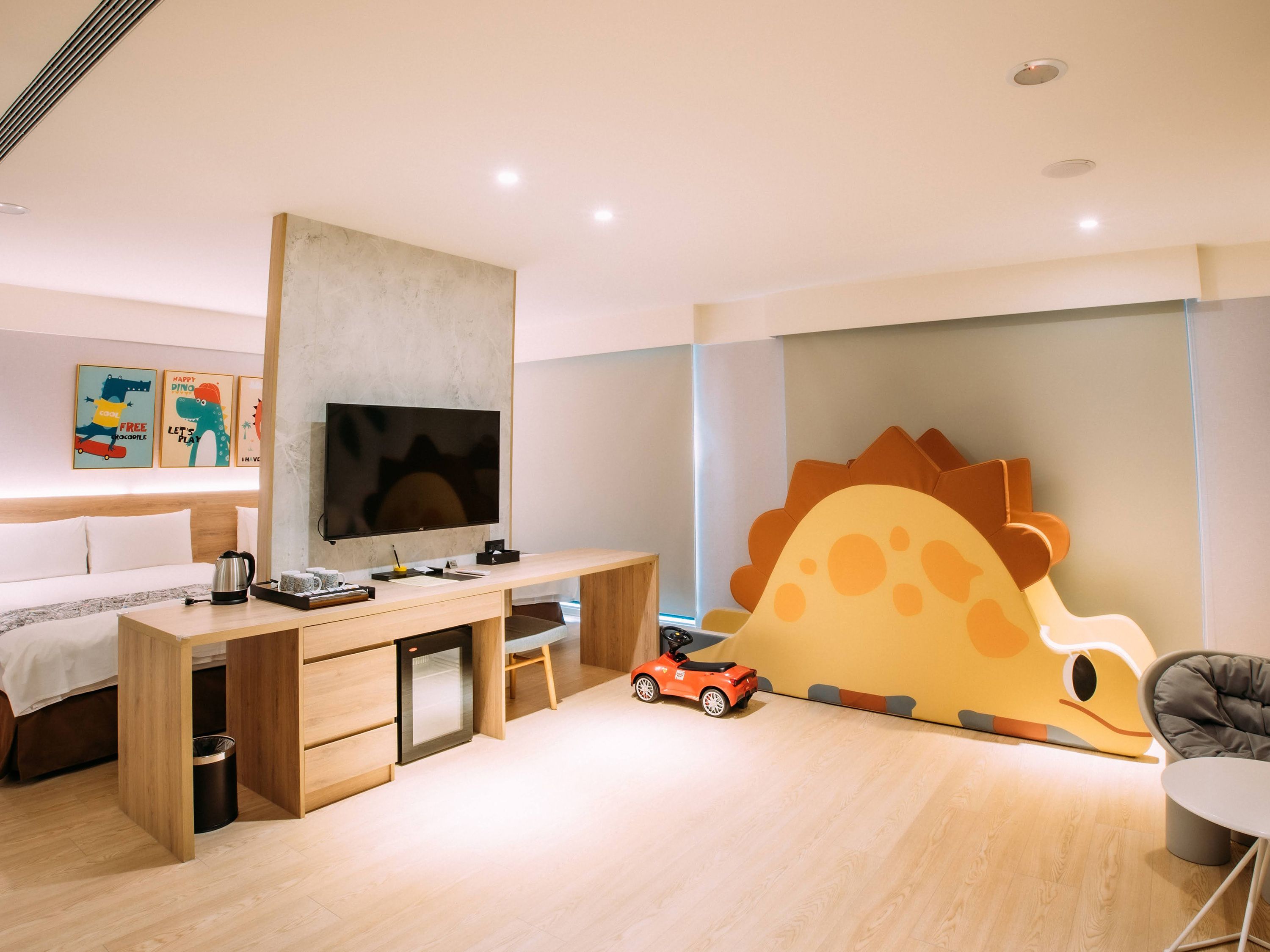 Una habitación de hotel moderna con un escritorio de madera, un televisor de pantalla plana, una decoración grande en la pared, un coche de juguete y una zona de estar cómoda.