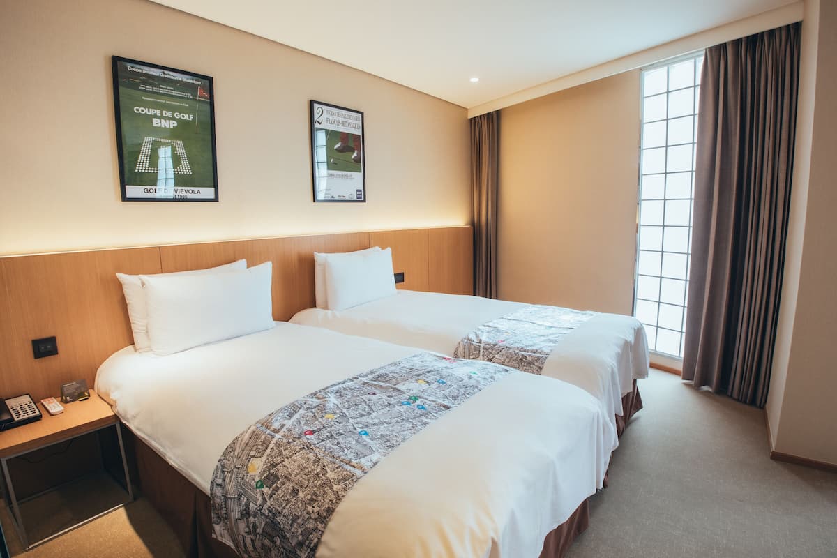 Habitación de hotel con dos camas, un cabecero de madera, una mesita de noche con teléfono y dos carteles enmarcados en la pared.