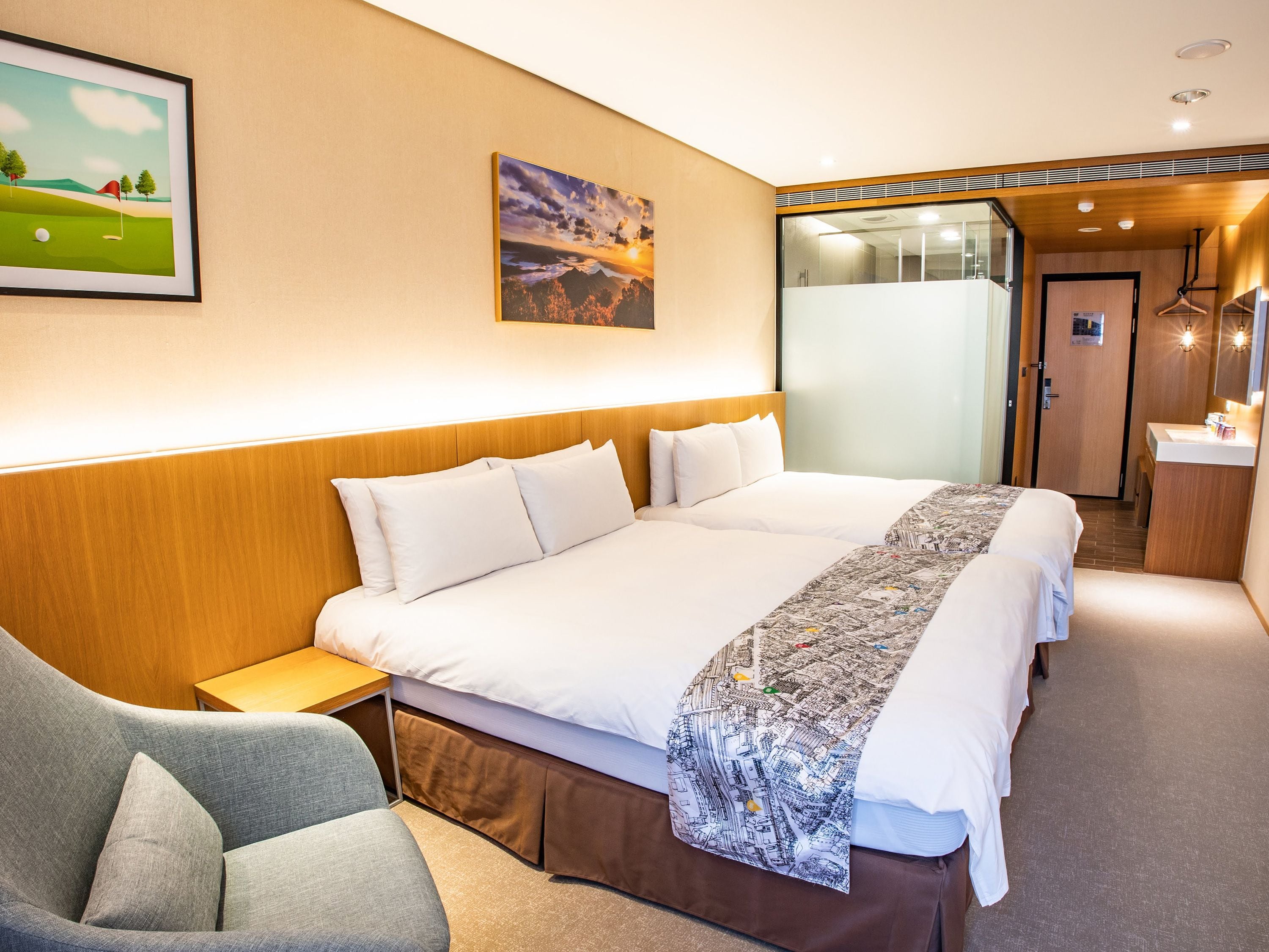 A hotel room with a large bed, a desk, a chair, a bathroom, and two paintings on the wall.